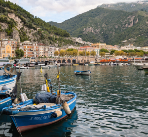 Kleine Fischerboote im Hafen einer italienischen K&uuml;stenstadt mit H&auml;usern am Hang und bewaldeten Bergen im Hintergrund.