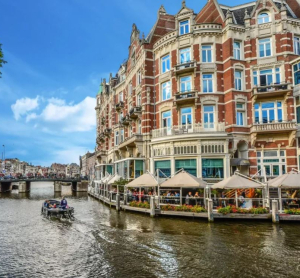 Historisches Geb&auml;ude am Kanal in Amsterdam mit Terrassencaf&eacute; und vorbeifahrendem Boot bei sonnigem Wetter.