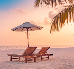 Zwei leere Liegestühle unter einem Sonnenschirm am Sandstrand bei Sonnenuntergang, mit Palmen im Vordergrund.