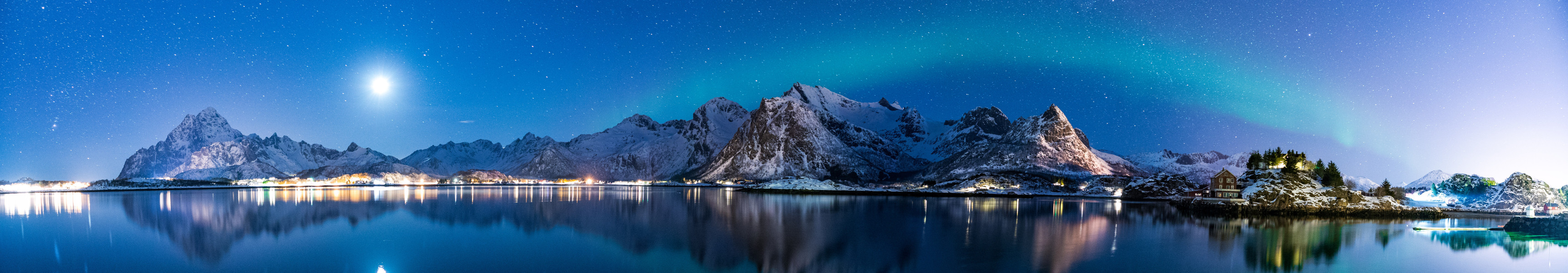 Panoramaaufnahme einer verschneiten Berglandschaft in Norwegen mit Nordlichtern und Spiegelung im ruhigen Wasser eines Fjords bei Nacht.