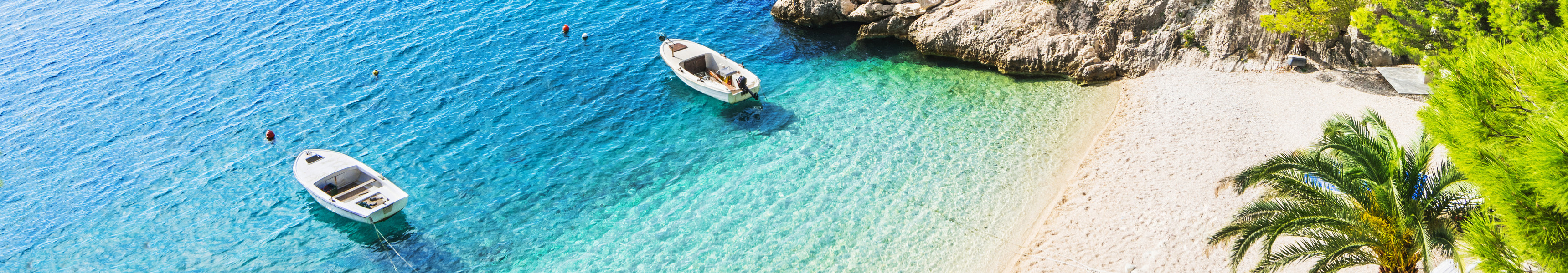Idyllischer Strand in Brela an der Makarska Riviera in Kroatien mit t&uuml;rkisfarbenem Wasser, wei&szlig;en Kieseln, kleinen Booten und Pinienb&auml;umen am Ufer.