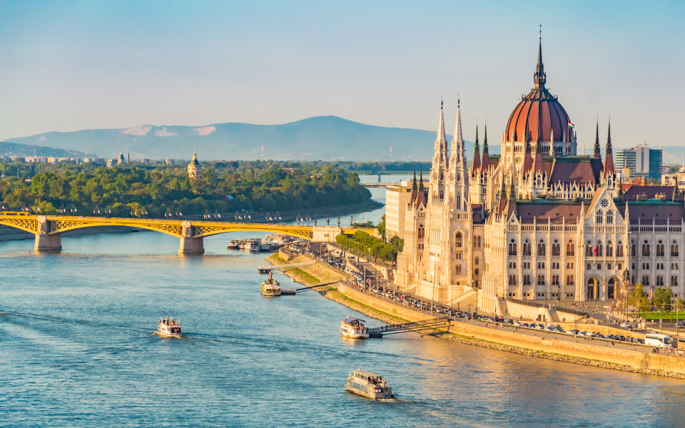 Ungarisches Parlamentsgebäude in Budapest, an der Donau mit Booten und einer Brücke im Hintergrund.