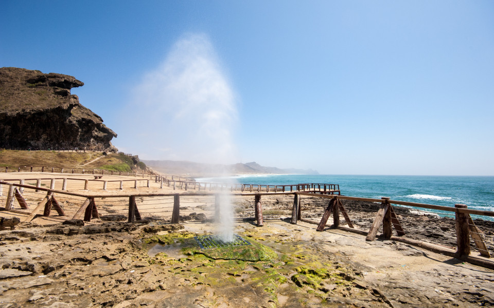 Al Mughsail Beach in Oman mit einem Wassergeysir, der aus den Felsen schie&szlig;t, vor dem weiten Ozean und einer Holzpromenade.
