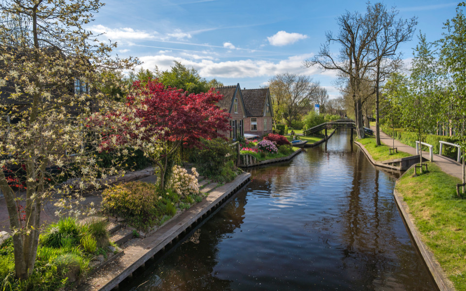 Kleines Backsteinhaus mit Garten an einem ruhigen Kanal in Giethoorn, umgeben von bl&uuml;henden B&auml;umen und Fu&szlig;g&auml;ngerbr&uuml;cken.