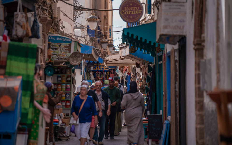 Belebte Gasse in der Altstadt von Essaouira mit Marktst&auml;nden, Schildern, bunten Gew&uuml;rzen und Touristen.